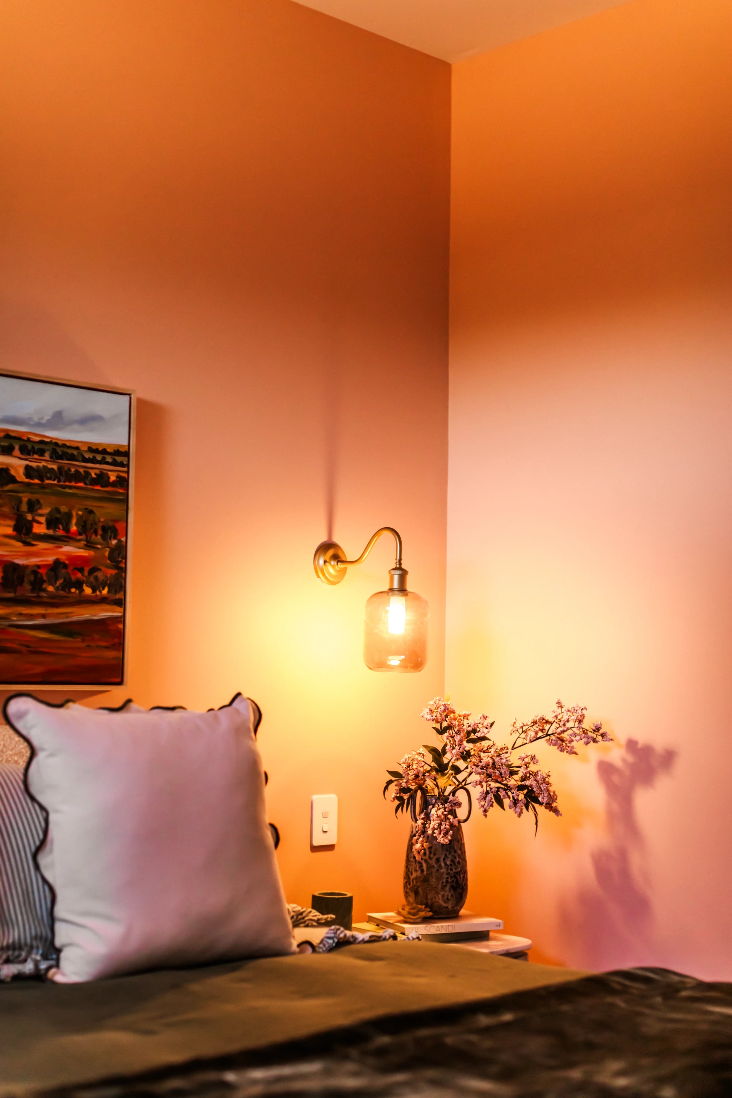 Cozy bedroom with orange walls, a bed with pillows, a lamp, and a vase with flowers.