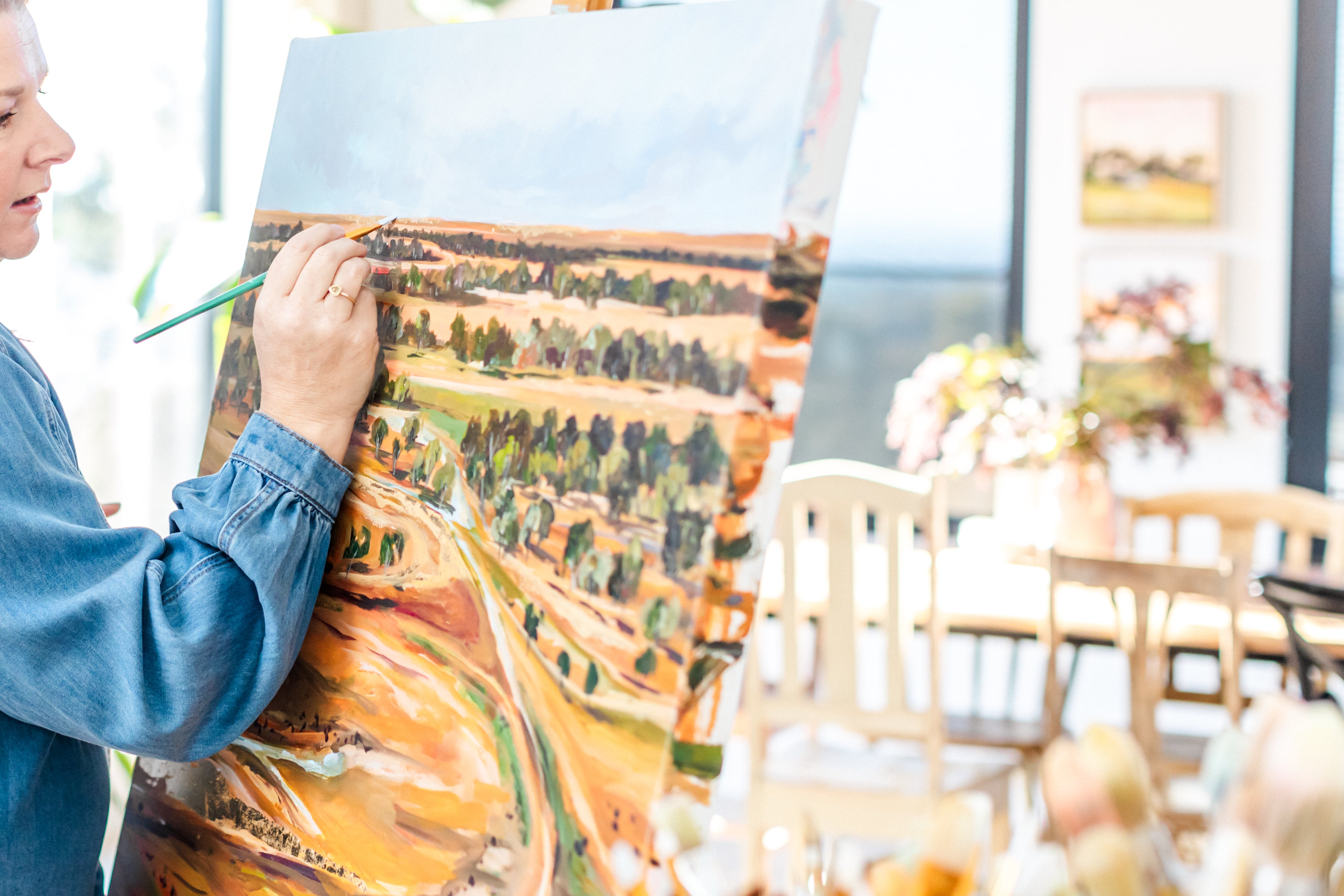 Person painting a landscape on an easel in a room with tables and chairs.
