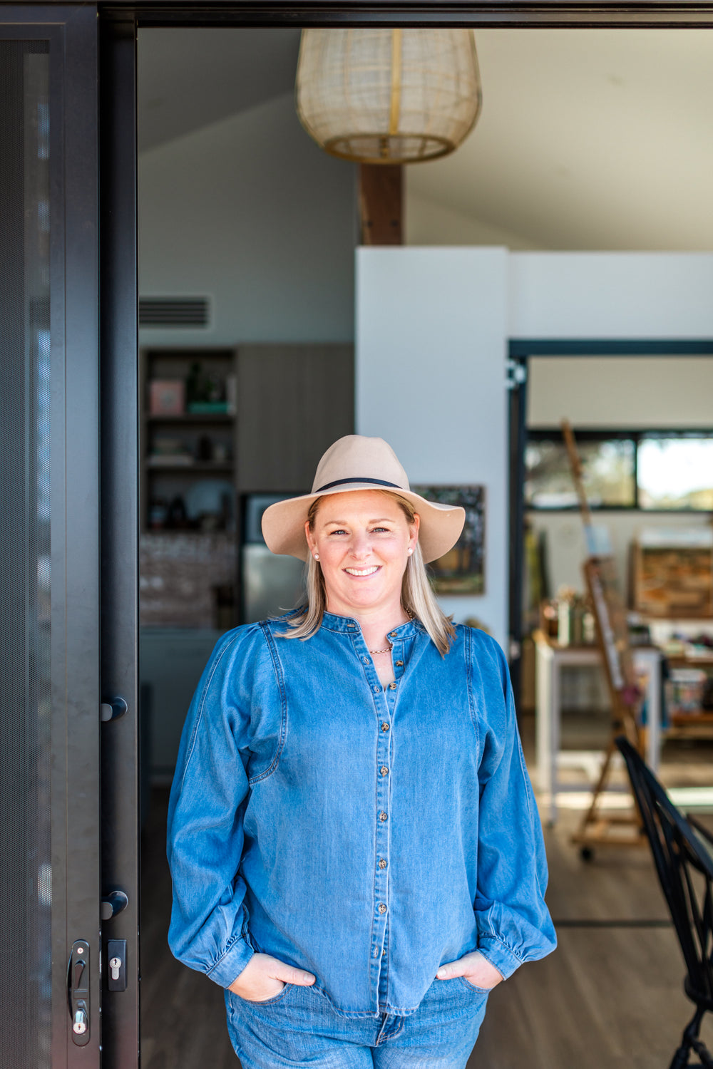 Woman wearing a blue denim outfit and beige hat standing in a room with a lamp and shelves.