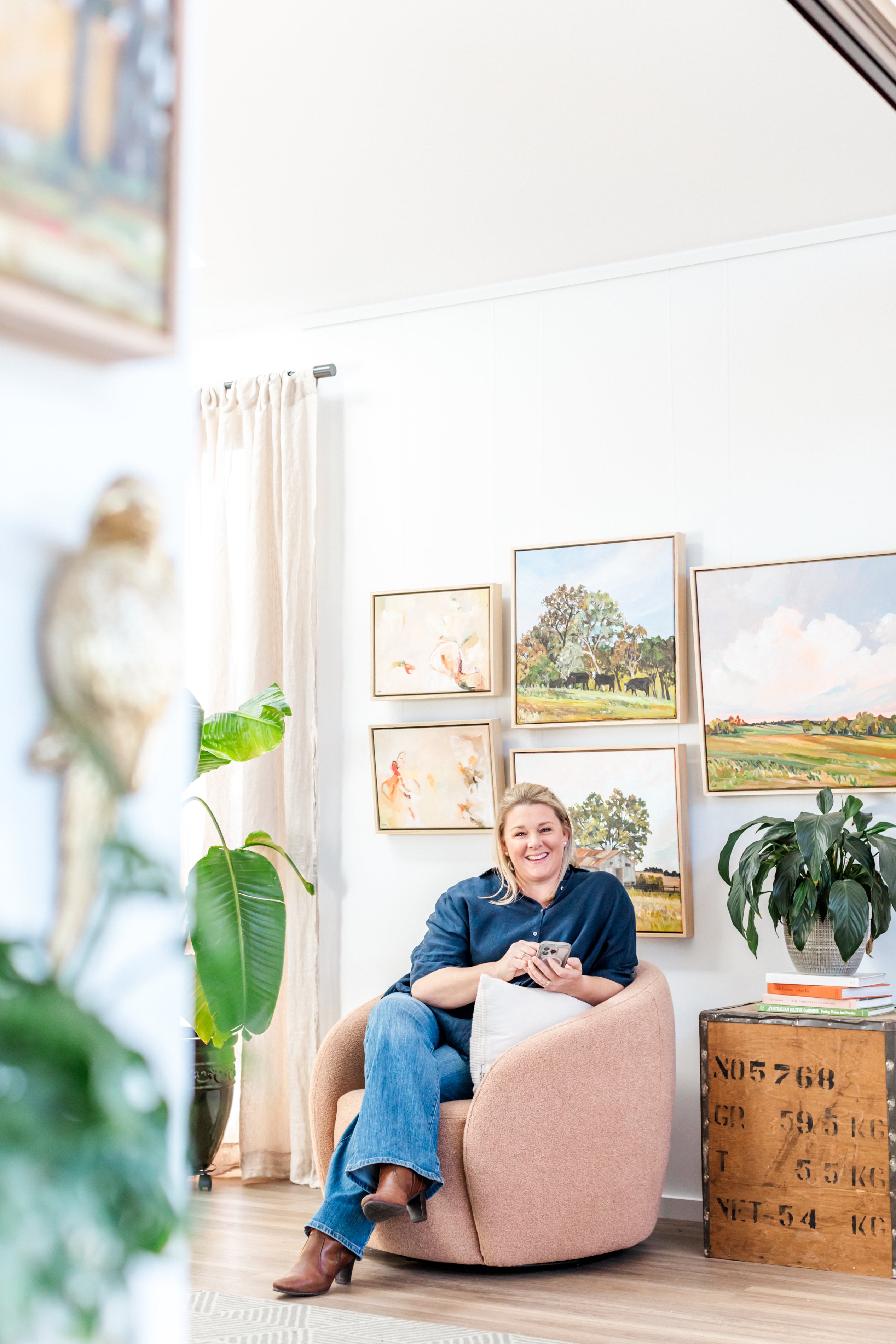 Woman sitting on a pink chair in a room with framed pictures and plants.
